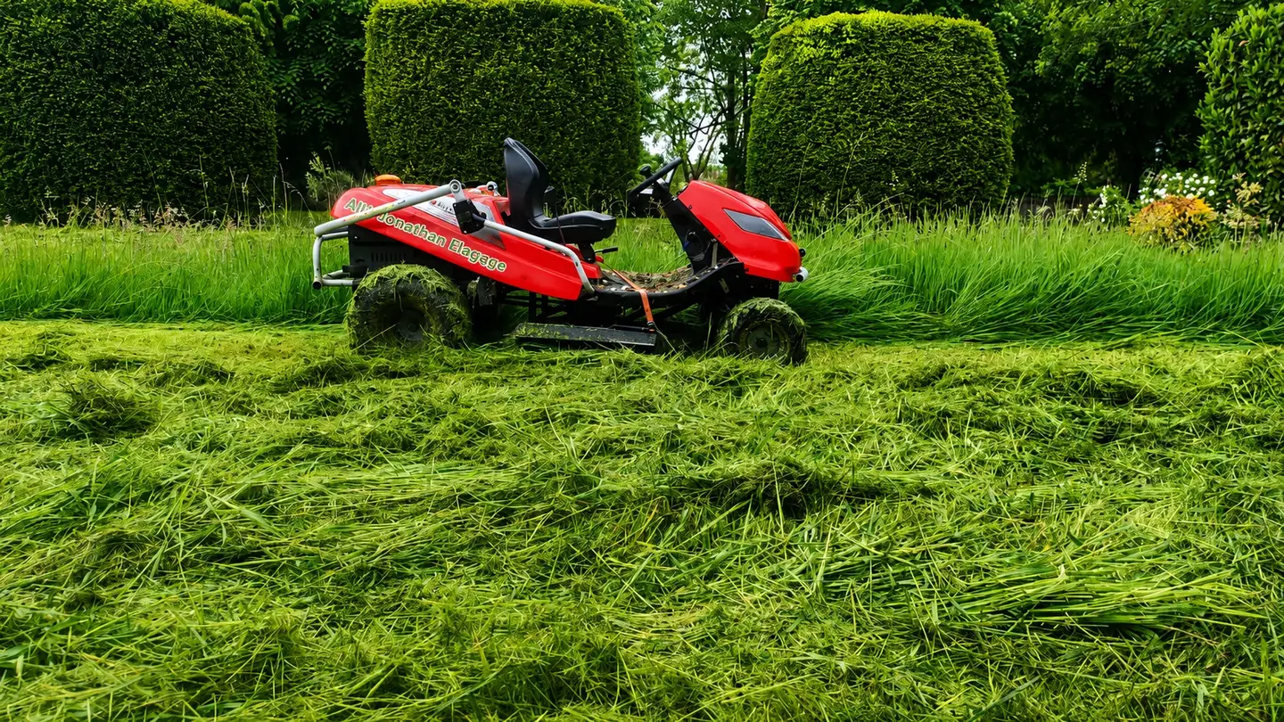 Tondeuse autoportée Allô Jonathan entre les haies sculptées d'un grand jardin, herbe haute fraîchement fauchée